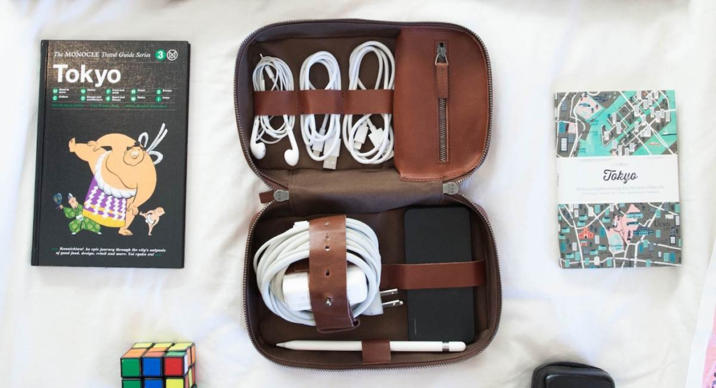 Flat lay of a travel guide, earphones, cords neatly stored in a This Is Ground Tech Dopp Kit, a notebook, map, and Rubik’s Cube on a white surface.