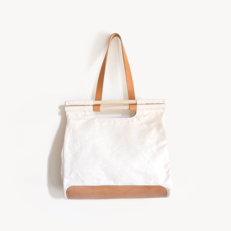 White canvas tote bag with tan leather handles and a leather bottom, hanging on a plain white wall.