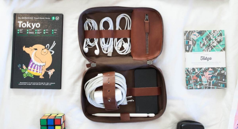 Flat lay of a travel guide, earphones, cords neatly stored in a This Is Ground Tech Dopp Kit, a notebook, map, and Rubik’s Cube on a white surface.